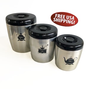 May include: Set of three silver canisters with black lids. The canisters have black and white illustrations of a teapot, coffee grinder, and sugar bowl. The canisters are labeled "Tea", "Coffee", and "Sugar".