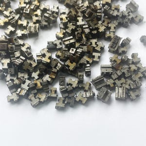 May include: A collection of small, metallic, T-shaped components scattered on a white surface. The components appear to be made of a dull, silver-colored metal with a slightly aged or weathered appearance. The shapes are uniform, suggesting they are manufactured parts.