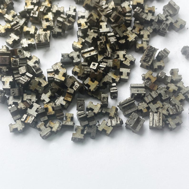 May include: A collection of small, metallic, T-shaped components scattered on a white surface. The components appear to be made of a dull, silver-colored metal with a slightly aged or weathered appearance. The shapes are uniform, suggesting they are manufactured parts.