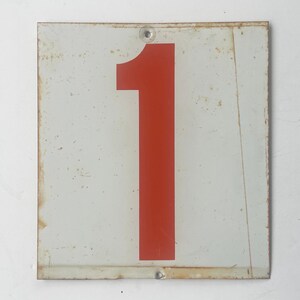 May include: Vintage metal sign featuring the number 1 in a bold red color against a white background. The sign has a weathered appearance with rust and wear around the edges, giving it a rustic, industrial aesthetic. The sign is square and has two holes for mounting.
