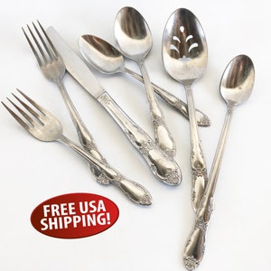 May include: Set of seven silver-toned flatware pieces, including a butter knife, a slotted spoon, a soup spoon, a teaspoon, and three forks. The handles have an ornate floral design.