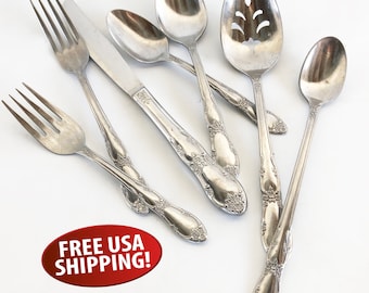 May include: Set of seven silver-toned flatware pieces, including a butter knife, a slotted spoon, a soup spoon, a teaspoon, and three forks. The handles have an ornate floral design.