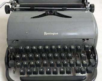 May include: A gray Remington Rand typewriter with a black keyboard and a carriage return lever. The typewriter has a black plastic case and a metal frame.