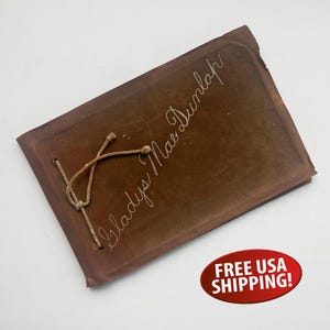 May include: A brown leather-bound photo album with a gold cord closure. The album is titled "Gladys Mae Dunlap" in white cursive writing.
