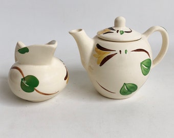 May include: A white ceramic teapot and creamer set with a brown and green floral design. The teapot has a lid and a handle. The creamer has a handle.