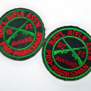 May include: Two black and green embroidered patches with red text. The patches are round and feature crossed rifles with the text "National Rifle Association" and "Pro-Marksman" around the outside. One patch says "15 ft. Award" and the other says "50 ft. Award".