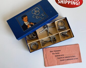 May include: A vintage puzzle set with a blue box featuring a man in a suit. The box contains several metal wire puzzles. A small booklet and a red oval sticker with the text "FREE USA SHIPPING!" are also visible.