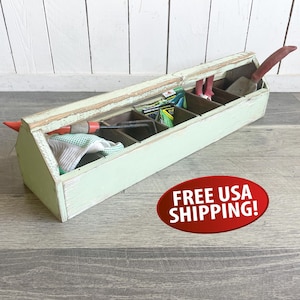May include: A distressed, light green wooden garden tool caddy with multiple compartments. The caddy contains gardening tools, seed packets, and a pair of gardening gloves. The caddy is on a wooden surface.