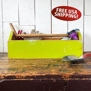 May include: A lime green wooden toolbox with a wooden handle, filled with gardening tools. The toolbox sits on a rustic wooden surface. Tools include a trowel, gloves, and a purple spray nozzle.