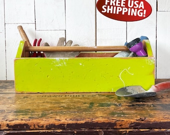 May include: A lime green wooden toolbox with a wooden handle, filled with gardening tools. The toolbox sits on a rustic wooden surface. Tools include a trowel, gloves, and a purple spray nozzle.