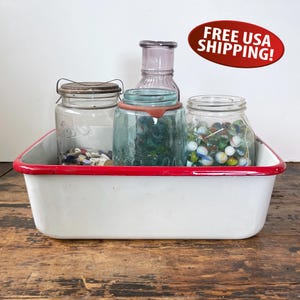 May include: A rectangular white enamel basin with a red rim containing three glass jars. One clear jar holds small buttons, a blue-tinted jar contains marbles, and a purple-tinted bottle stands behind them. The basin has a smooth, glossy finish.