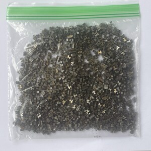 May include: A clear plastic bag filled with numerous small, metallic-looking, geometric shapes. The shapes appear to be a dark, muted silver color, with some having a slightly golden hue. The bag has a green zipper closure.