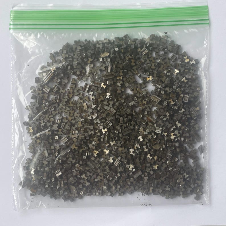 May include: A clear plastic bag filled with numerous small, metallic-looking, geometric shapes. The shapes appear to be a dark, muted silver color, with some having a slightly golden hue. The bag has a green zipper closure.