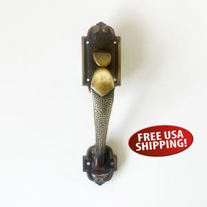 May include: Ornate bronze door handle with a textured grip and decorative backplate. The handle features a unique design with a curved top and a detailed, patterned grip, ideal for vintage or antique doors.