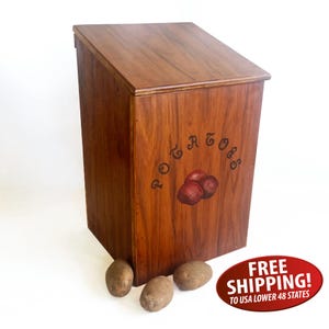 May include: A wooden potato bin with a slanted lid and a dark brown finish. The bin features the word "Potatoes" in a curved design, along with an illustration of red potatoes. Three potatoes are at the base of the bin.