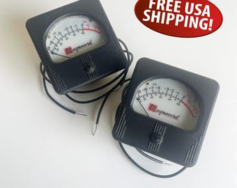 May include: Two vintage black Magnecord VU meters with white faces, black needles, and red accents. Each meter has a curved scale and a small knob. Black wires extend from the meters.
