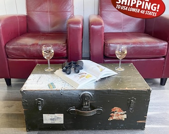 May include: Two deep red leather armchairs positioned behind a rectangular black wooden trunk. The trunk features a central metal latch, a handle, and two white labels. On top sits an open book, a pair of black binoculars, and two wine glasses.