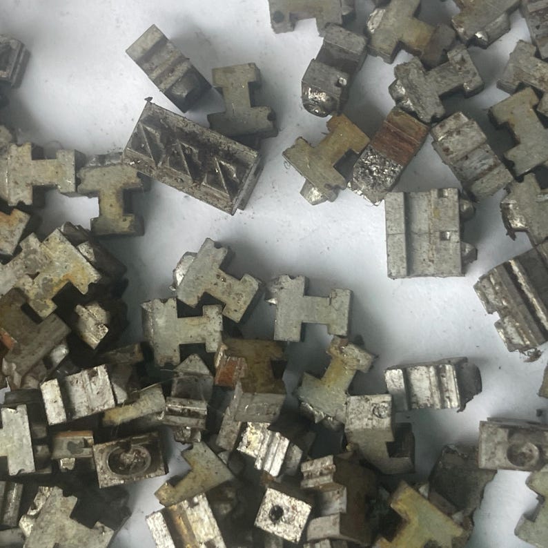 May include: A collection of small, weathered, metallic pieces, many shaped like the letter "T" or rectangular blocks. Some pieces show signs of wear and age, with a dull, gray color. The objects are scattered across a light-colored surface.