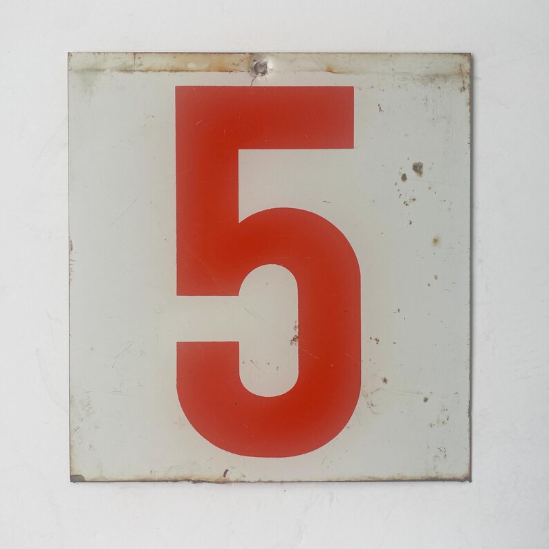 May include: A square, vintage metal sign with a white background and a large, bold red number 5. The sign has a slightly distressed appearance, with minor wear and tear visible. A small nail secures it to the wall.