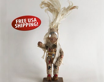 May include: A vintage doll wearing a white feathered headdress and a white and brown patterned outfit. The doll is standing on a wooden base.