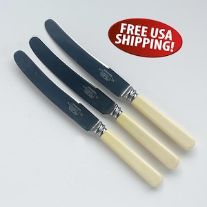 May include: Three stainless steel knives with cream-colored handles. The blades are dark and shiny, with text that reads "STAINLESS S. PEARSON & CO SHEFFIELD ENGLAND". The knives are arranged diagonally on a white surface.