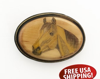 May include: A wooden belt buckle with a hand-painted portrait of a brown horse's head. The buckle is oval-shaped and has a black metal frame.