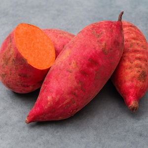 May include: Close-up of several sweet potatoes with reddish-purple skin. One is cut in half, revealing bright orange flesh. The other sweet potatoes are whole, with a slightly rough texture and a tapered shape. The background is a solid gray.