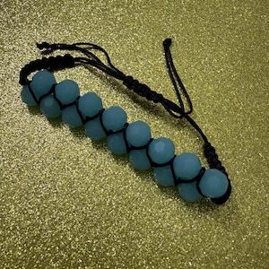 Op de afbeelding: Handgemaakte armband met lichtblauwe facetkralen op zwart koord. De armband heeft een verstelbare macramé sluiting. De kralen hebben een zachte, pastelkleur. Het ontwerp is eenvoudig en elegant.