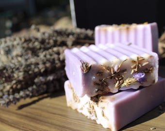 May include: Two lavender-colored soap bars are stacked, adorned with dried lavender and small purple flowers. The top bar has a ridged surface. Dried lavender sprigs are in the background, with a wooden surface.