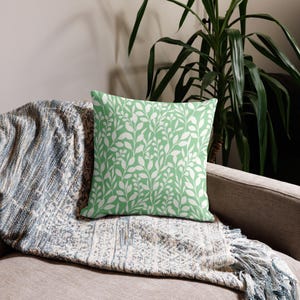 May include: A square cushion featuring a light green backdrop and a white leaf design. It rests on a grey sofa, covered with a blue and white patterned throw. A large green plant is visible in the background.