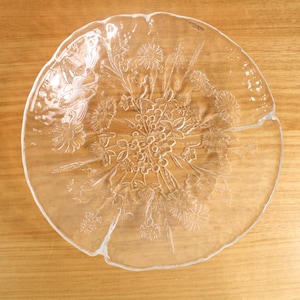 May include: A clear glass serving plate with an irregular, organic edge and a textured, embossed floral pattern featuring various wildflowers and stems across the surface. The brand name Kosta Boda is etched in small letters near the top rim.