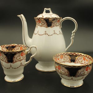 May include: A white porcelain tea set comprising a teapot, creamer jug, and sugar bowl. The set has an ornate design with orange, navy, and gold detailing. The teapot features a curved spout and handle.