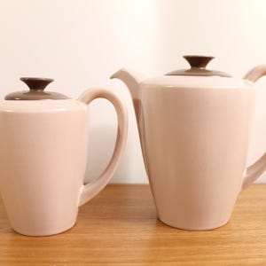 May include: Two ceramic teapots in a soft pale pink finish with smooth, tapered bodies and curved handles. Each teapot features a distinct, dark brown circular lid with a small knob handle at the top and a short, angled spout for pouring.