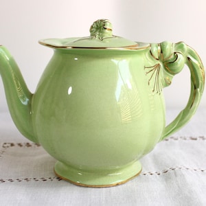 May include: A pale green ceramic teapot with a lid and a curved handle. The teapot has gold trim around the base, lid, and handle. The handle has a decorative bow design. The teapot is on a white surface.