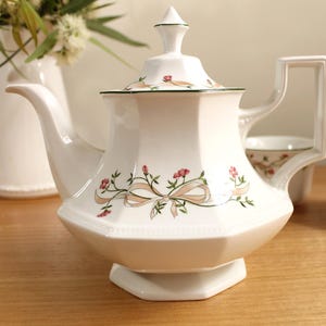 May include: White porcelain teapot with a floral and ribbon design. The teapot has a lid with a pointed finial and a square handle. The design includes pink flowers and a tan ribbon. The teapot is on a wooden surface.