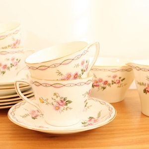 May include: A set of cream-coloured porcelain teacups, saucers, a creamer, and a sugar bowl. Each piece features a delicate pink floral pattern, green leaves, and a thin pink scalloped border with gold trim along the edges of the ceramic items.