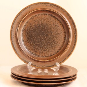 May include: Four brown ceramic plates with a speckled design. The plates have a wide rim and a dark brown centre. One plate is displayed on a clear plastic stand. The plates are likely for serving food, perhaps at a dinner party.