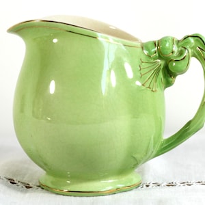May include: A pale green ceramic jug with a rounded body and a curved handle. The jug has gold trim around the rim, base, and handle details. The handle features a decorative floral design.