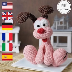 Puede incluir: Un perro rosa tejido a mano con orejas marrones y una nariz roja. El perro tiene ojos negros y está sentado en un plato blanco. La imagen incluye una etiqueta de "PDF pattern".