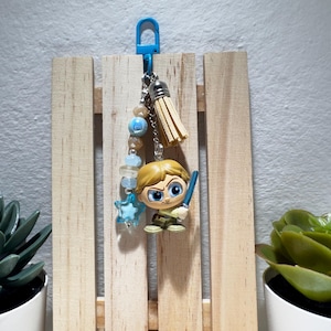 May include: A keychain with a cartoon character holding a lightsaber, suspended from a blue clip. The charm has a blonde head and is wearing a tan outfit. The keychain includes a beige tassel and blue and clear beads.
