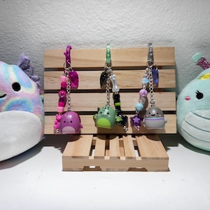 May include: Three keychains with charms of various creatures and a wooden display stand. The keychains are attached to a wooden display. Two plush toys are on either side of the display. One is a purple and blue axolotl, and the other is a teal creature with a floral crown.