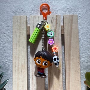 Könnte beinhalten: Ein Schlüsselanhänger mit einer Cartoonfigur mit schwarzen Haaren, die ein rotes Hemd und eine blaue Hose trägt. Der Schlüsselanhänger enthält eine grüne Quaste, eine Kette und bunte Blumen- und Totenkopfperlen. Er hat einen orangefarbenen Clip.
