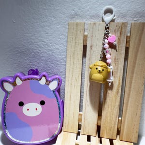 以下が含まれることがあります： 紫とピンクの牛の形をした容器で、キラキラした縁取りがあります。 黄色のクマのキーホルダーにはビーズチェーンとハートのチャームが付いており、木の板の背景にぶら下がっています。 アイテムは白い表面にあります。