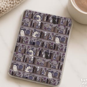 Può includere: Una custodia trasparente per telefono con un motivo ripetuto di una libreria con fantasmi, gatti, candele e bottiglie di pozioni. Lo sfondo è un viola tenue con accenti floreali. Una tazza di caffè e un maglione beige sono sullo sfondo.
