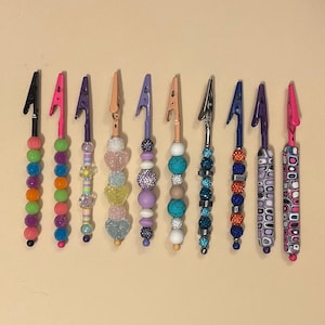 May include: A collection of ten decorative alligator clips with colorful beaded accents. Each clip features a different design, including various colors of beads, rhinestones, and patterns. The clips are arranged in a row against a neutral background.