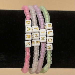 May include: Four beaded bracelets in shades of pink, purple, and green. The bracelets spell out "DARK THE IN GLOW" with white letter beads. The beads are strung on clear elastic.