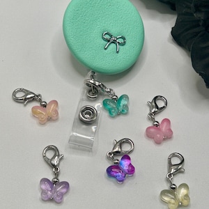 May include: A mint green circular badge reel featuring a small silver bow emblem, accompanied by six individual butterfly charms in translucent shades of pink, teal, purple, and yellow, each attached to a silver-toned metal lobster clasp.