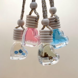 May include: Heart-shaped car air fresheners hanging from gray cords. The clear glass bottles contain colorful liquids and decorative elements. The tops are light gray with wooden beads. One bottle has blue flowers, another has pink liquid, and another has blue glitter.