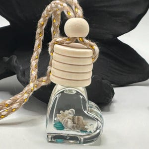 May include: A heart-shaped glass bottle filled with small seashells and turquoise stones. The bottle features a light-colored wooden cap with horizontal ridges and a round wooden topper, suspended by a braided beige rope with gold metallic threads.