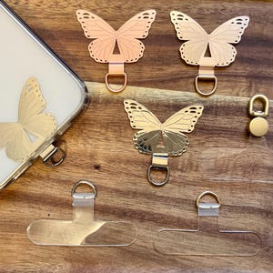 Pode incluir: Uma coleção de pingentes de telefone em forma de borboleta em tons de ouro, ouro rosa e prata, juntamente com abas transparentes para aderência ao telefone. Um pingente está preso a uma capa de telefone. Os pingentes têm um laço para fixação.